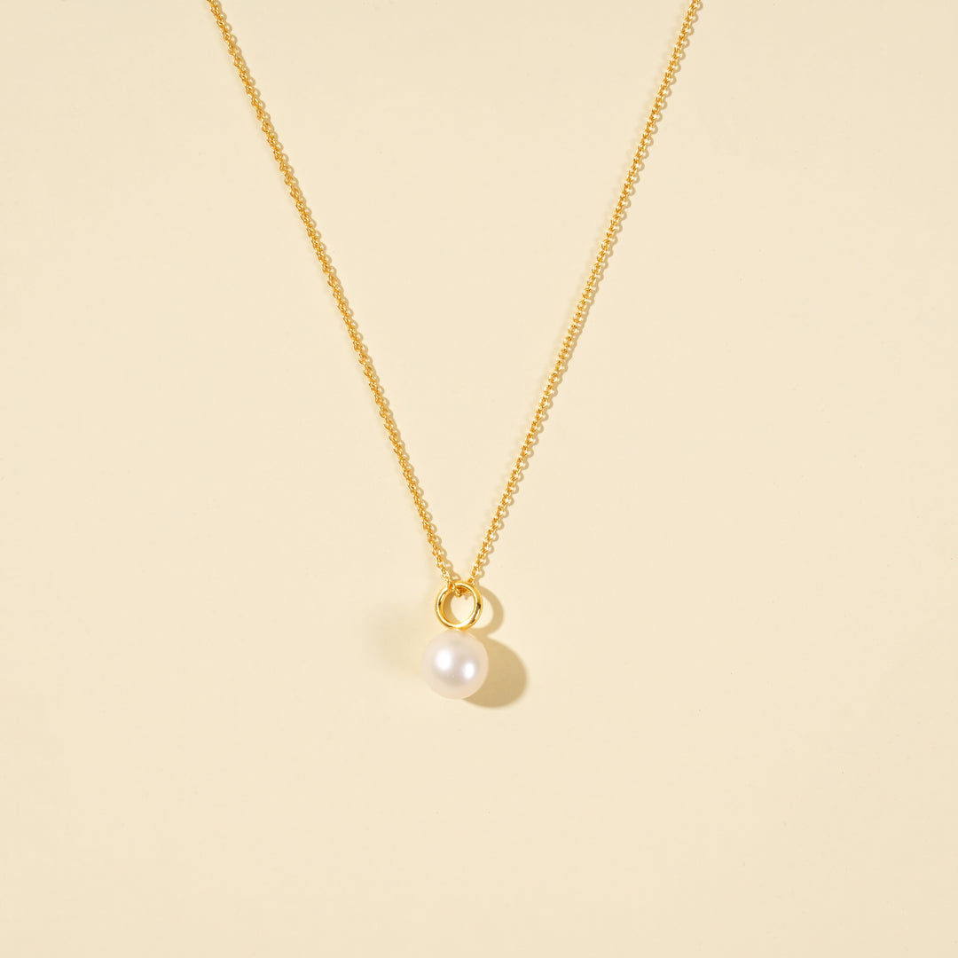 A clean, minimalist product shot of a gold pearl necklace on a neutral cream background. The image clearly displays the pearl necklace with cable chain.