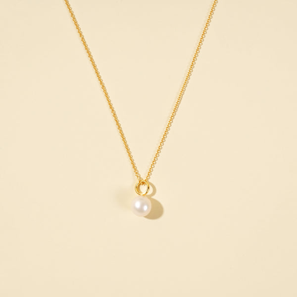 A clean, minimalist product shot of a gold pearl necklace on a neutral cream background. The image clearly displays the pearl necklace with cable chain.