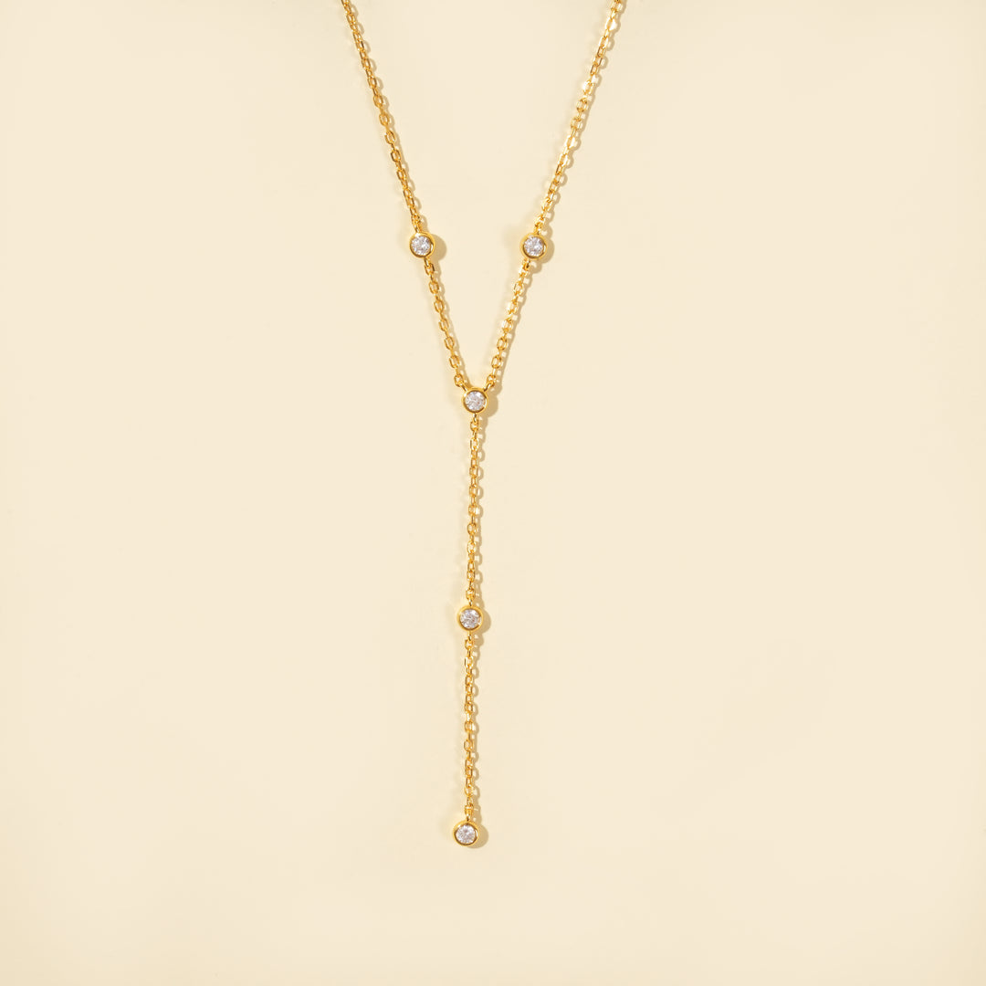 A clean, minimalist product shot of a gold lariat necklace on a neutral cream background. The image clearly displays the delicate Y-shape and the five sparkling, bezel-set crystals that adorn the chain and drop.