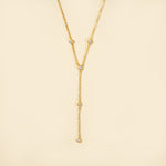 Load image into Gallery viewer, A clean, minimalist product shot of a gold lariat necklace on a neutral cream background. The image clearly displays the delicate Y-shape and the five sparkling, bezel-set crystals that adorn the chain and drop.
