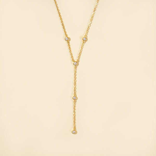 A clean, minimalist product shot of a gold lariat necklace on a neutral cream background. The image clearly displays the delicate Y-shape and the five sparkling, bezel-set crystals that adorn the chain and drop.