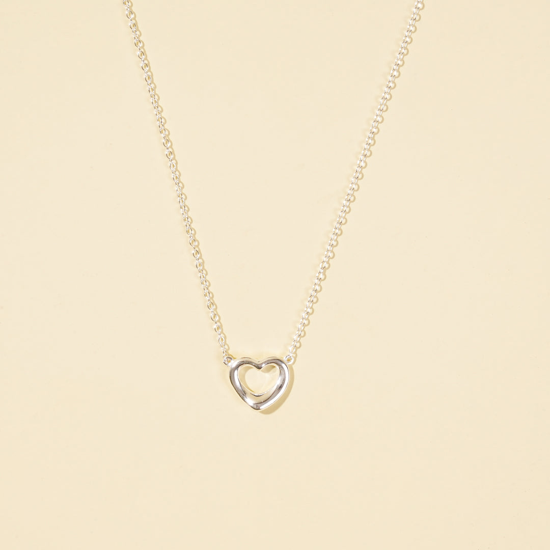  A clean, minimalist product shot of a silver open heart necklace on a neutral cream background. The image clearly displays the delicate chain and the simple, polished open heart pendant, a classic and timeless piece of jewellery.