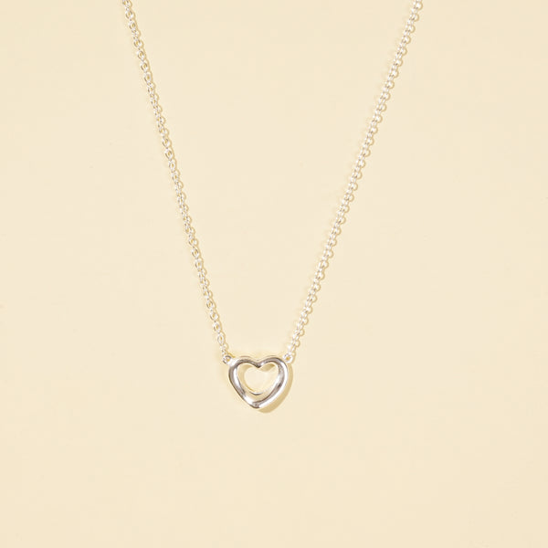  A clean, minimalist product shot of a silver open heart necklace on a neutral cream background. The image clearly displays the delicate chain and the simple, polished open heart pendant, a classic and timeless piece of jewellery.