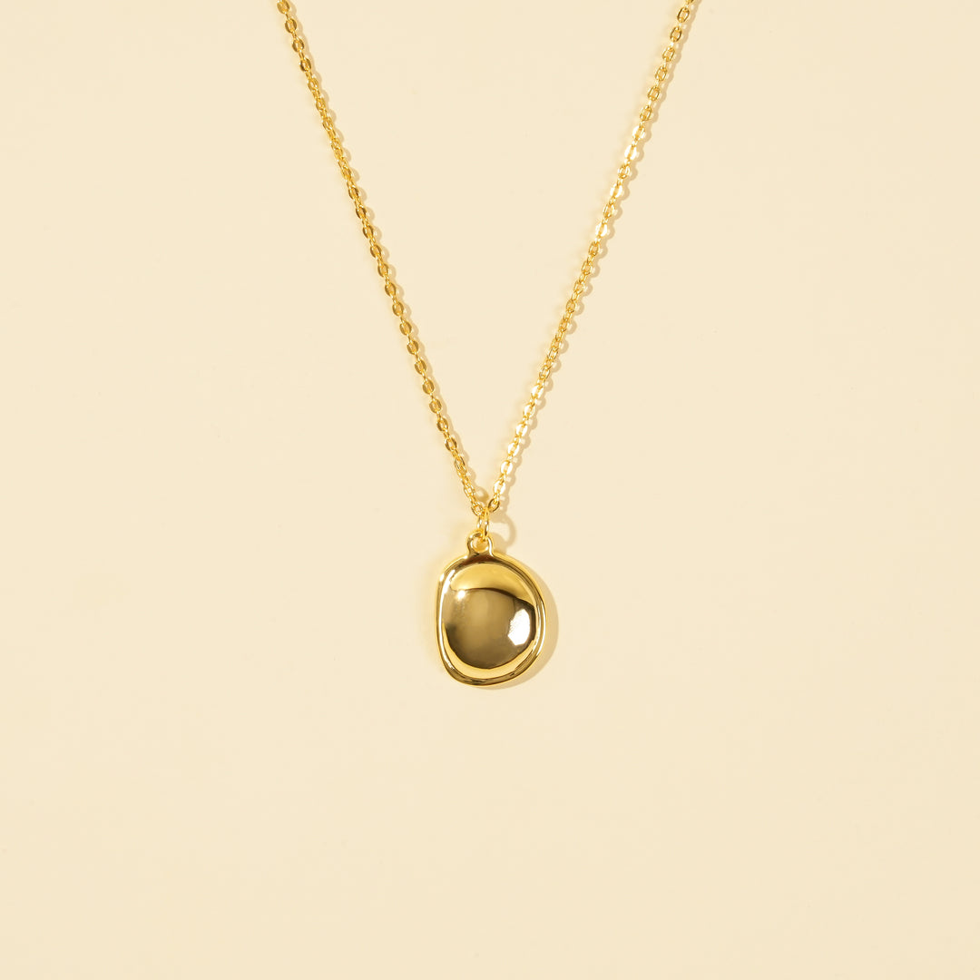 A clean, minimalist product shot of a gold necklace with an organically shaped, bezel-set stone pendant. The image clearly displays the smoky translucent gemstone and delicate gold chain against a neutral cream background.