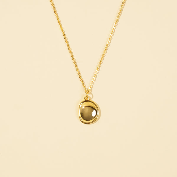 A clean, minimalist product shot of a gold necklace with an organically shaped, bezel-set stone pendant. The image clearly displays the smoky translucent gemstone and delicate gold chain against a neutral cream background.