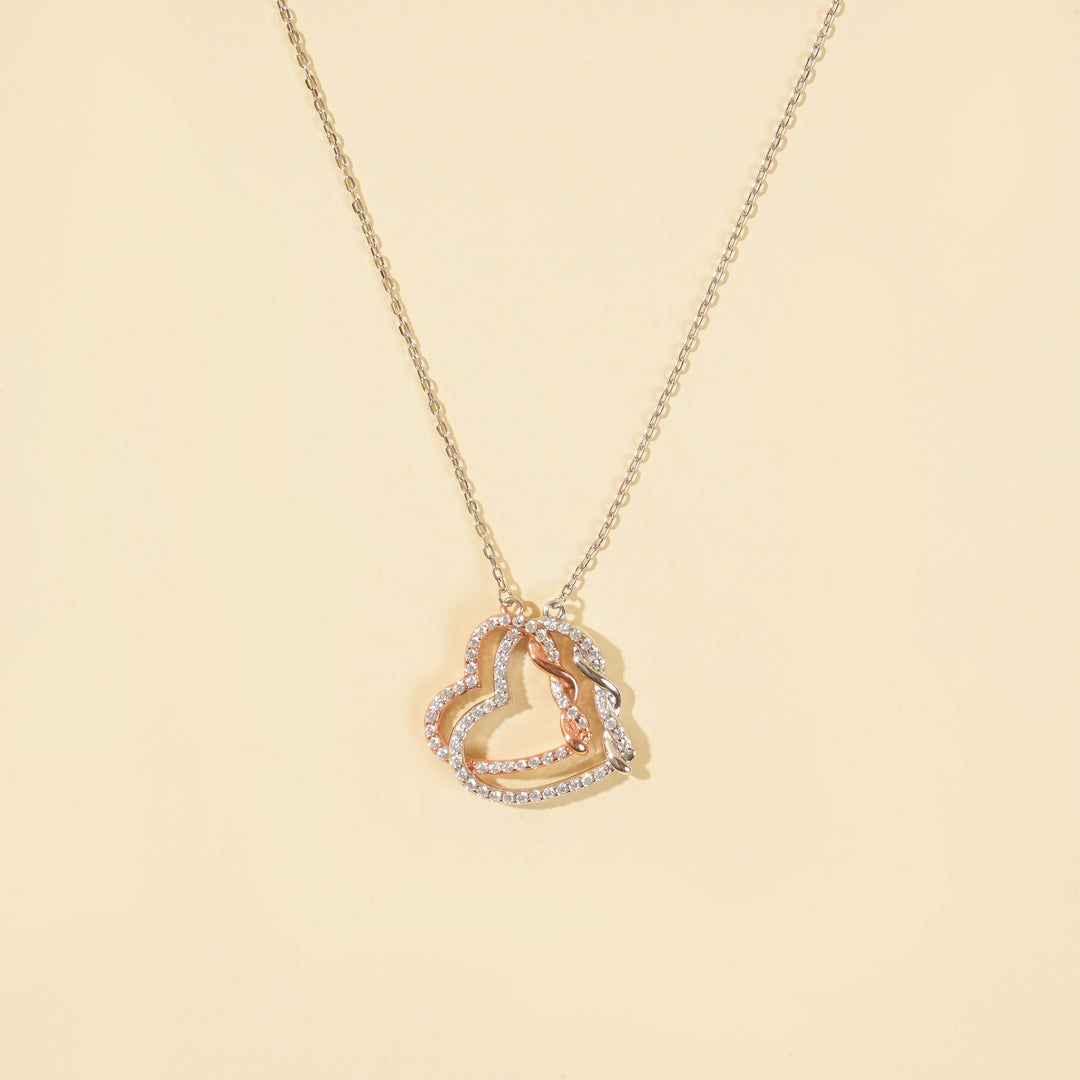 A clean product shot of a two-tone interlocking hearts necklace on a neutral cream background. The image clearly displays the intricate silver and rose gold pavé crystal hearts, a beautiful symbol of love and connection.