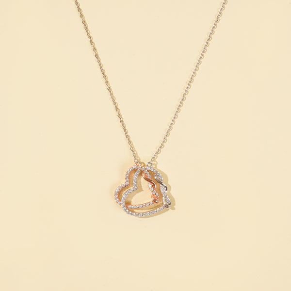 A clean product shot of a two-tone interlocking hearts necklace on a neutral cream background. The image clearly displays the intricate silver and rose gold pavé crystal hearts, a beautiful symbol of love and connection.