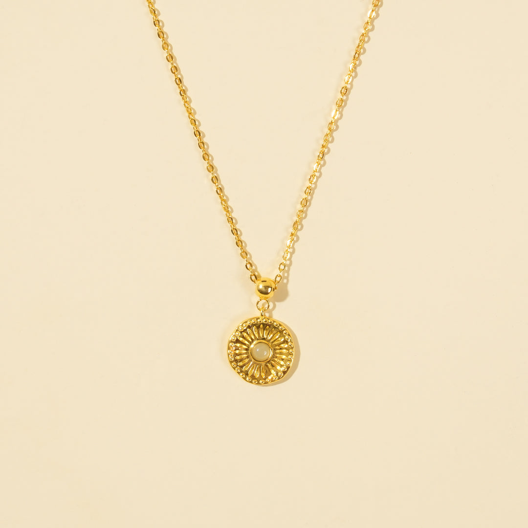 A clean, minimalist product shot of a gold sun medallion necklace on a neutral cream background. The image clearly displays the intricate sunburst texture, central opalite stone, and beaded border of the celestial-themed pendant.