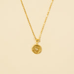 Load image into Gallery viewer, A clean, minimalist product shot of a gold sun medallion necklace on a neutral cream background. The image clearly displays the intricate sunburst texture, central opalite stone, and beaded border of the celestial-themed pendant.
