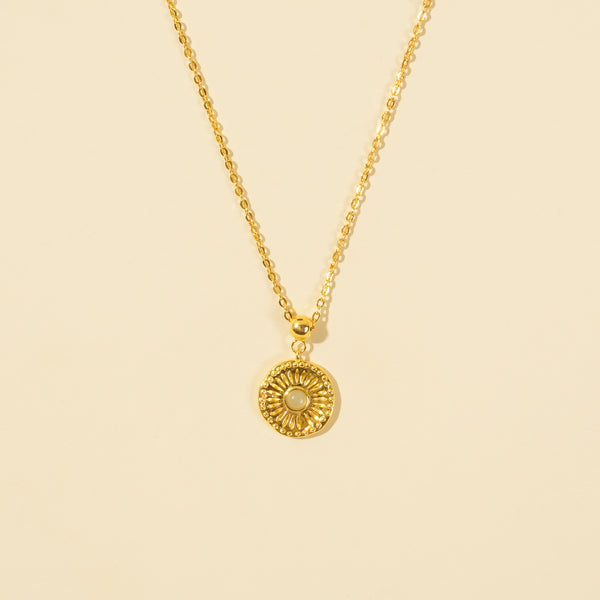A clean, minimalist product shot of a gold sun medallion necklace on a neutral cream background. The image clearly displays the intricate sunburst texture, central opalite stone, and beaded border of the celestial-themed pendant.