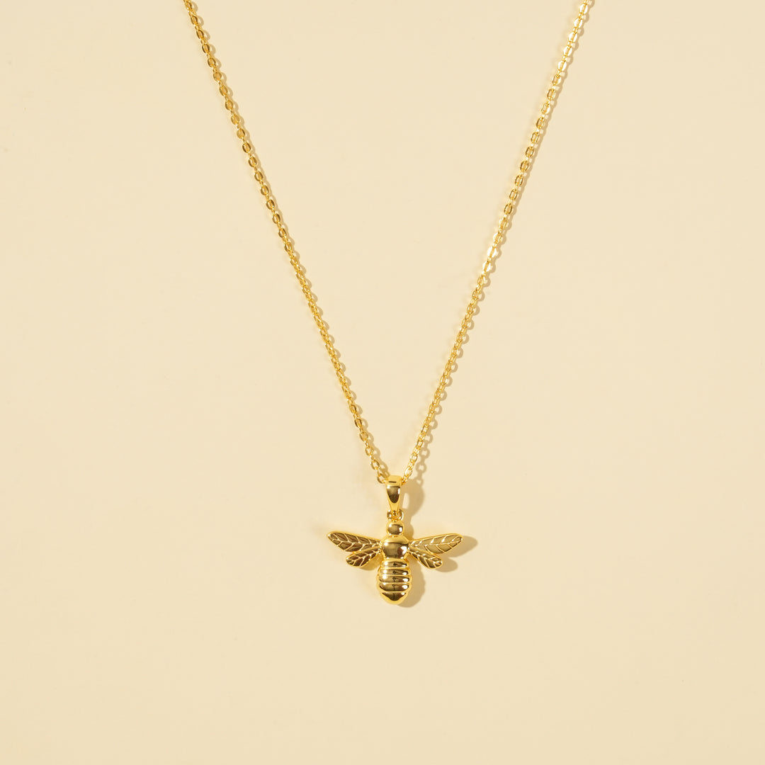 A clean, minimalist product shot of a gold bumblebee necklace on a neutral background. The image clearly displays the intricate details of the bee pendant and the delicate gold chain, a perfect piece of nature-inspired jewellery.