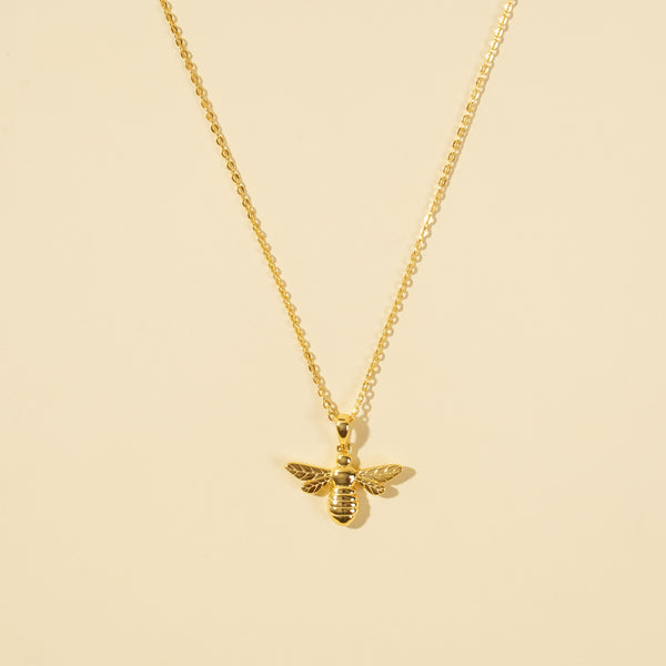 A clean, minimalist product shot of a gold bumblebee necklace on a neutral background. The image clearly displays the intricate details of the bee pendant and the delicate gold chain, a perfect piece of nature-inspired jewellery.