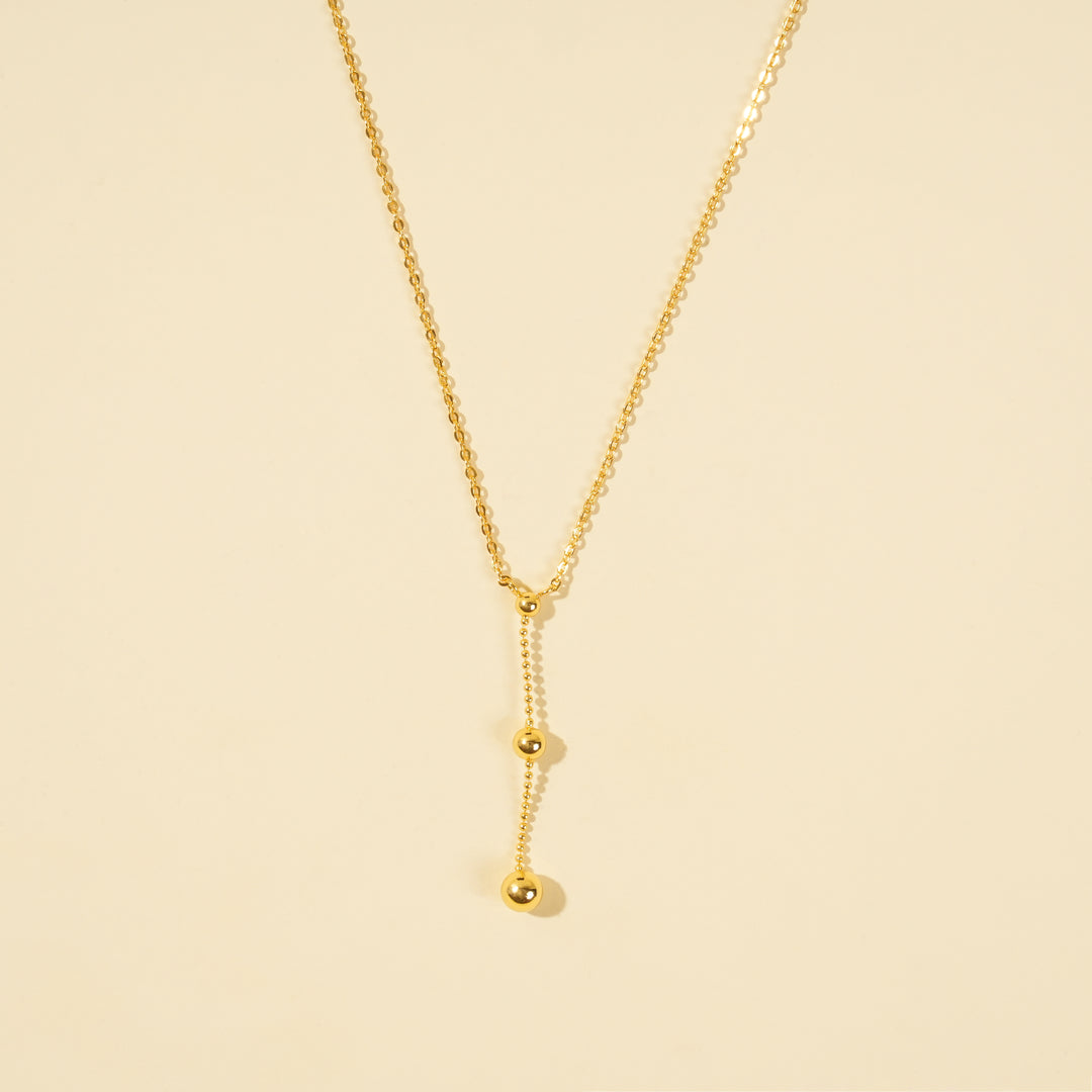 A clean, minimalist product shot of a gold Y-necklace on a neutral cream background. The image clearly displays the lariat design with a delicate drop chain accented by two polished gold spheres of graduating sizes.