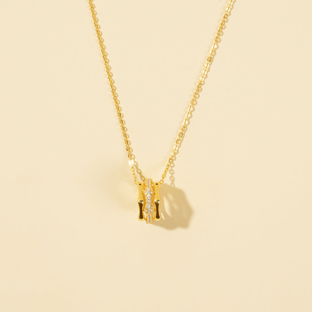 A clean, minimalist product shot of a modern gold necklace on a neutral background. The image clearly shows the cylindrical pendant with its polished gold bands and a sparkling central row of pavé-set crystals.