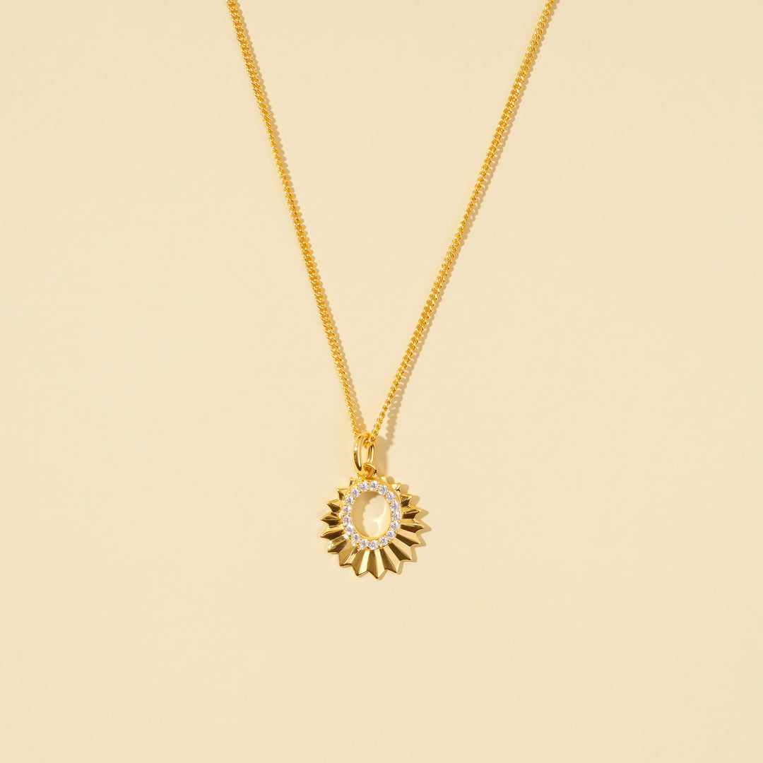 A clean, minimalist product shot of a gold celestial sun necklace on a neutral cream background. The image clearly displays the faceted sun-ray design and the open oval center lined with sparkling pavé crystals.