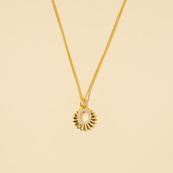 A clean, minimalist product shot of a gold celestial sun necklace on a neutral cream background. The image clearly displays the faceted sun-ray design and the open oval center lined with sparkling pavé crystals.