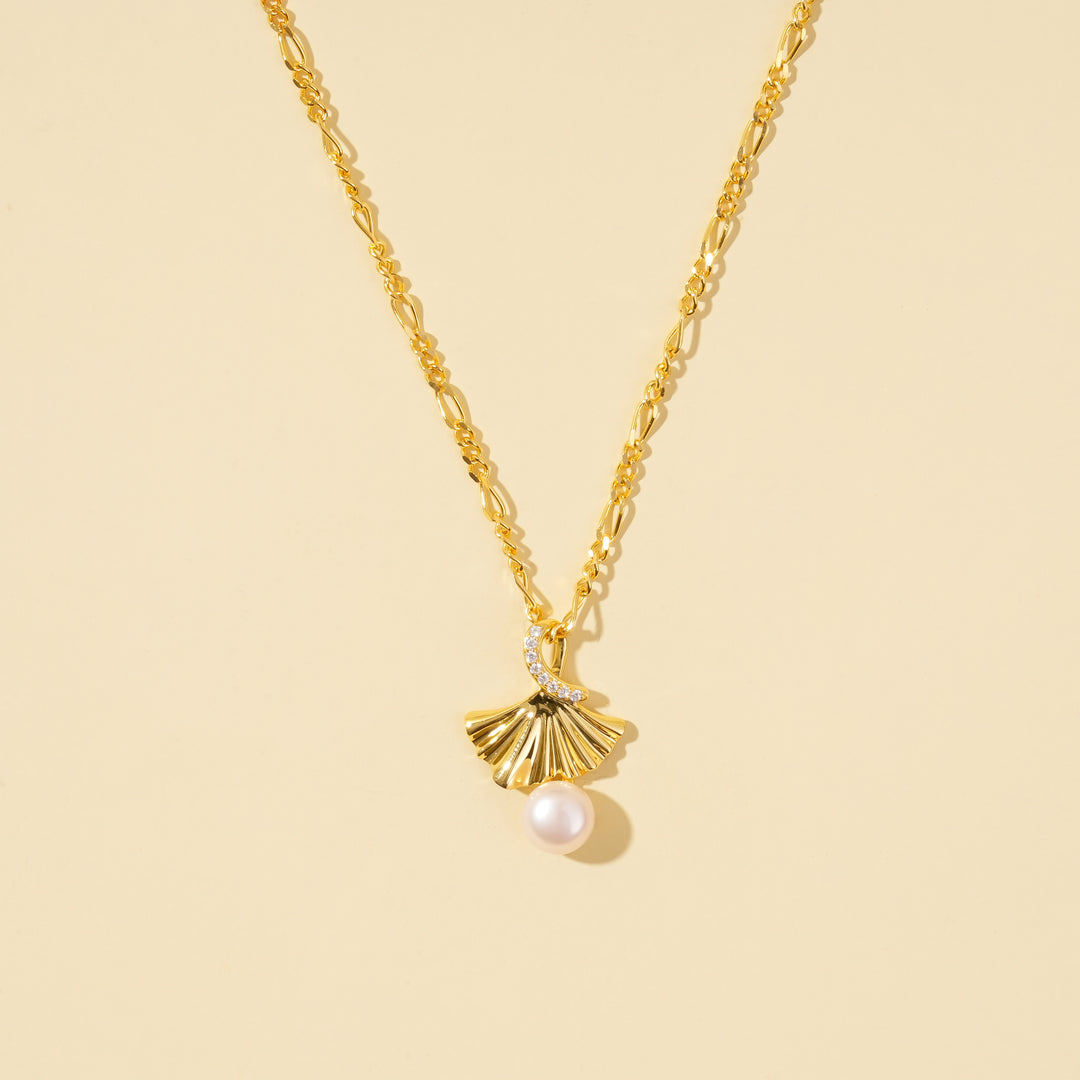  clean product shot of a gold ginkgo leaf and pearl necklace on a neutral cream background. The image clearly displays the pleated, fan-shaped pendant with its dangling pearl, crystal accent, and decorative link chain.