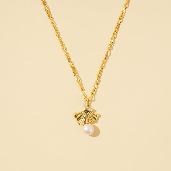  clean product shot of a gold ginkgo leaf and pearl necklace on a neutral cream background. The image clearly displays the pleated, fan-shaped pendant with its dangling pearl, crystal accent, and decorative link chain.