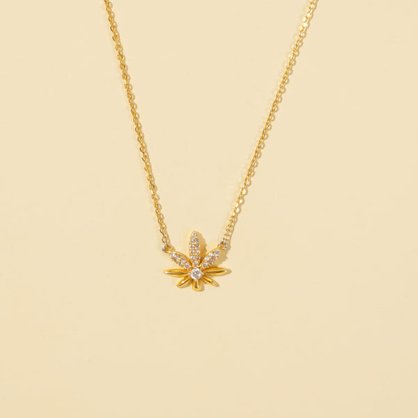 A clean, minimalist product shot of a gold cannabis leaf necklace on a neutral cream background. The image clearly displays the intricate pavé crystal detailing and the central solitaire stone on the trendy pot leaf pendant.