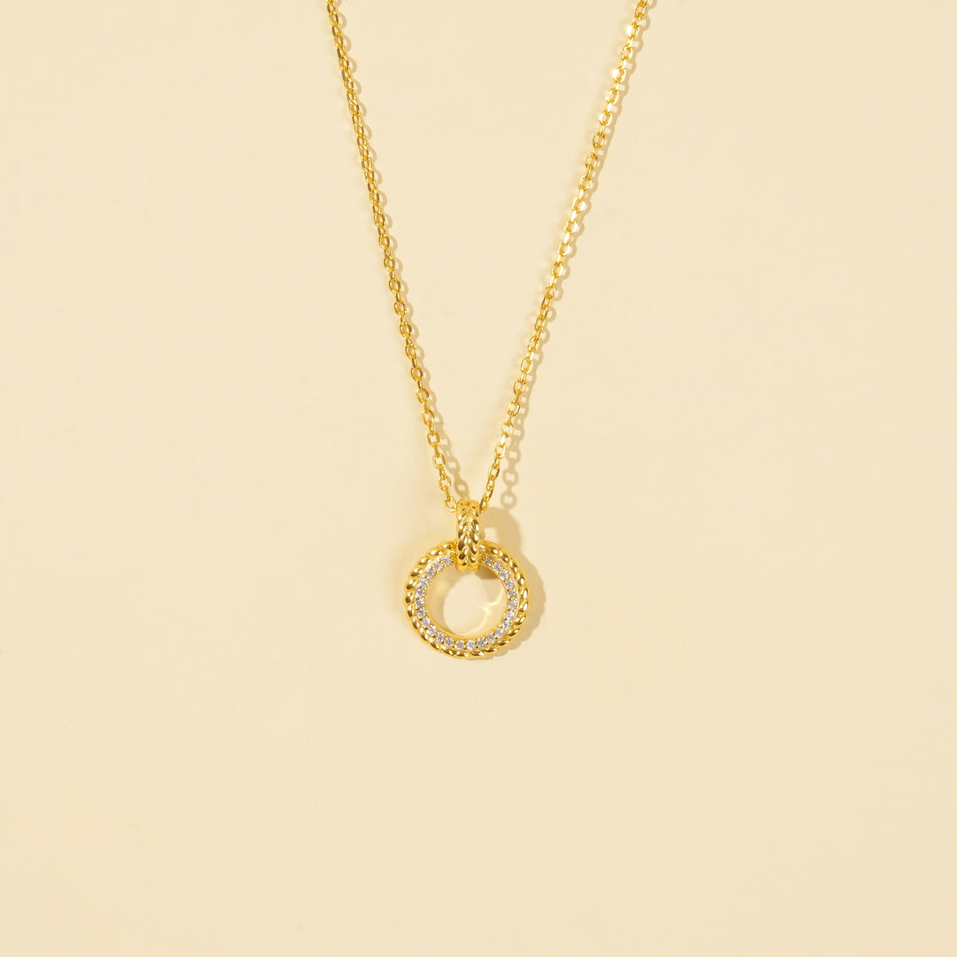 A clean, minimalist product shot of a gold circle pendant necklace on a neutral cream background. The image clearly displays the intricate design, with one half of the ring featuring sparkling pavé crystals and the other half a polished, twisted rope texture.