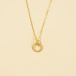 Load image into Gallery viewer, A clean, minimalist product shot of a gold circle pendant necklace on a neutral cream background. The image clearly displays the intricate design, with one half of the ring featuring sparkling pavé crystals and the other half a polished, twisted rope texture.
