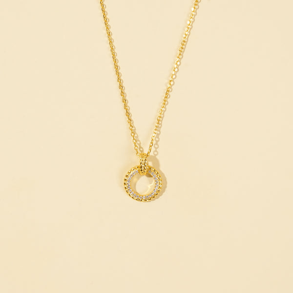 A clean, minimalist product shot of a gold circle pendant necklace on a neutral cream background. The image clearly displays the intricate design, with one half of the ring featuring sparkling pavé crystals and the other half a polished, twisted rope texture.
