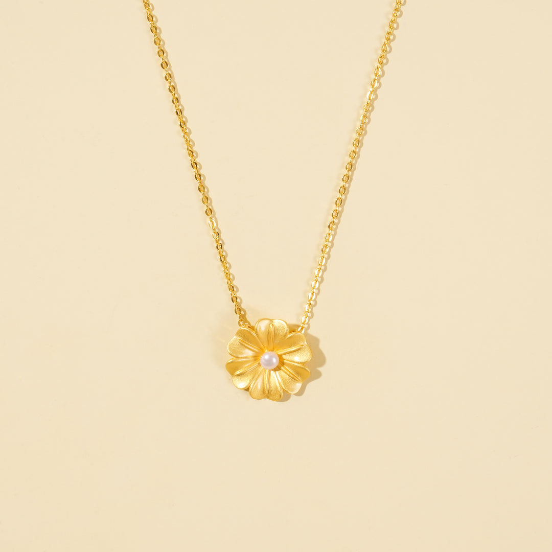 A clean, minimalist product shot of a gold floral necklace on a neutral background. The image highlights the textured petals and single pearl at the center of the flower pendant, showcasing its delicate botanical design.