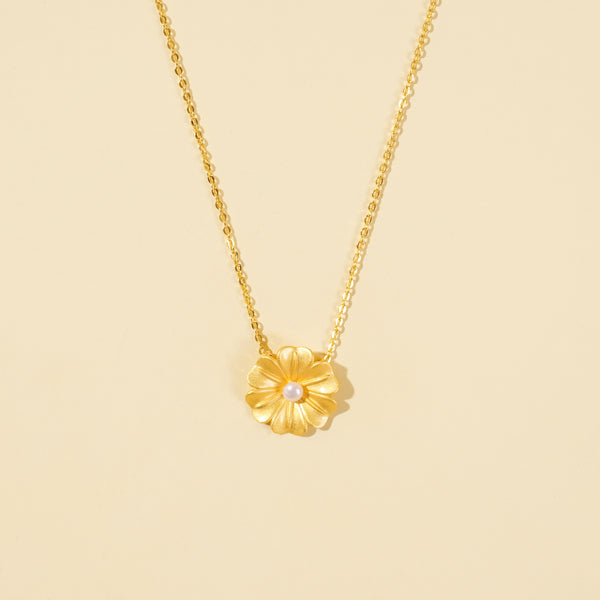 A clean, minimalist product shot of a gold floral necklace on a neutral background. The image highlights the textured petals and single pearl at the center of the flower pendant, showcasing its delicate botanical design.