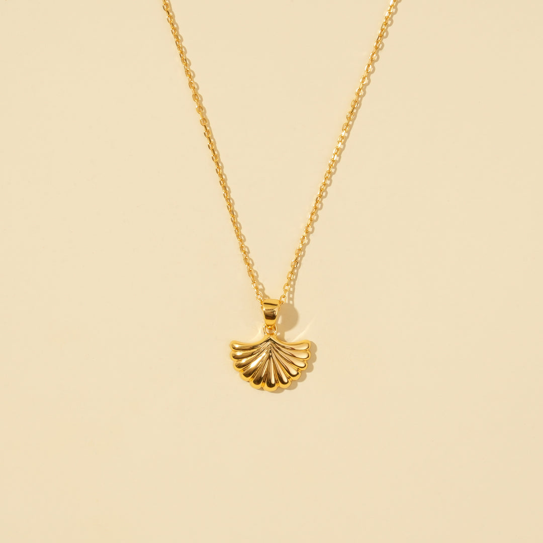 A clean, minimalist product shot of a gold seashell necklace on a neutral cream background. The image clearly displays the detailed, ribbed texture of the scallop shell pendant and its delicate chain.