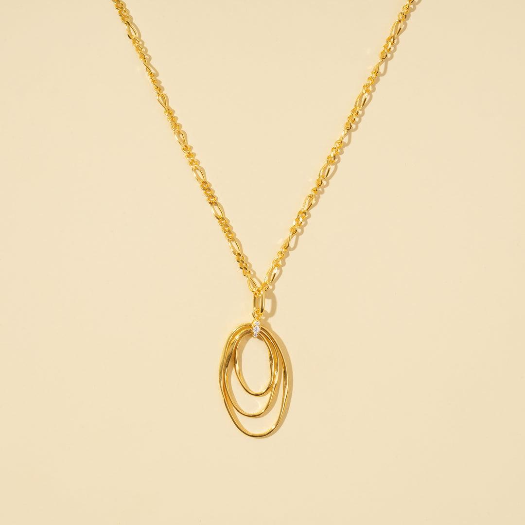A clean, minimalist product shot of a gold necklace featuring a triple interlocking oval pendant with a crystal accent. The contemporary design and elegant figaro chain are clearly displayed on a neutral cream background.