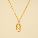 Load image into Gallery viewer, A clean, minimalist product shot of a gold necklace featuring a triple interlocking oval pendant with a crystal accent. The contemporary design and elegant figaro chain are clearly displayed on a neutral cream background.
