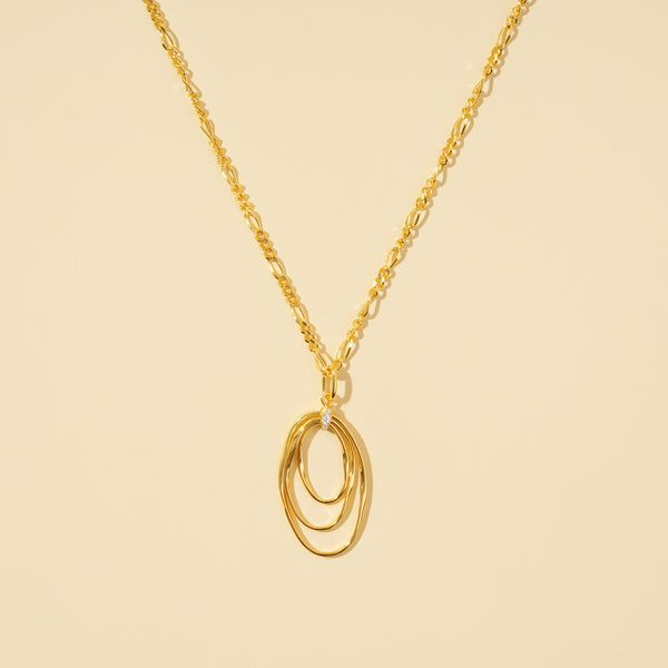 A clean, minimalist product shot of a gold necklace featuring a triple interlocking oval pendant with a crystal accent. The contemporary design and elegant figaro chain are clearly displayed on a neutral cream background.