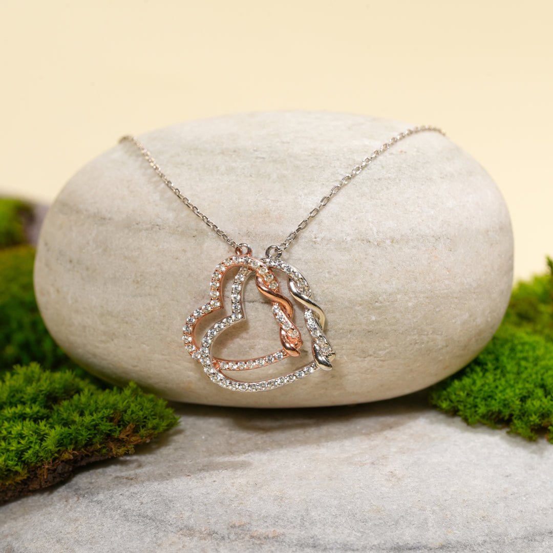 An elegant two-tone double heart necklace from Zewory Jewellery, displayed in a serene natural setting. The sparkling, crystal-encrusted silver and rose gold hearts are draped over a smooth, light-grey stone and surrounded by fresh green moss.