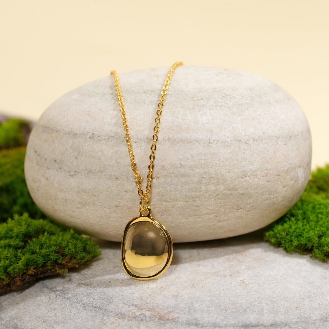 A modern gold gemstone necklace from Zewory Jewellery, displayed in a serene natural setting. The unique, pebble-shaped smoky stone pendant is draped over a smooth, light-grey stone and surrounded by fresh green moss.