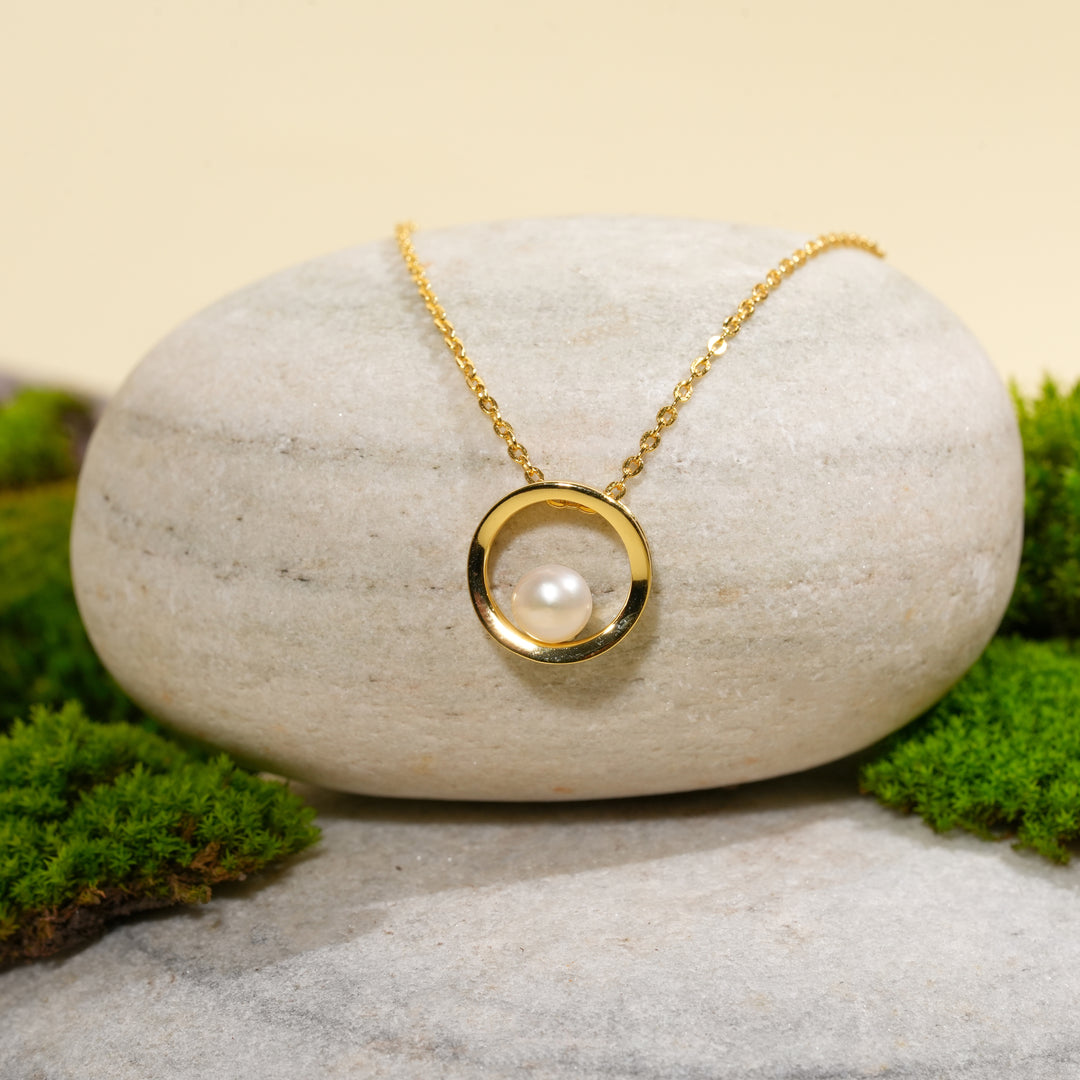 A contemporary gold necklace from Zewory Jewellery, featuring an open circle pendant with a central pearl. The elegant, minimalist piece is displayed on a smooth, light-grey stone and surrounded by fresh green moss for a natural aesthetic.