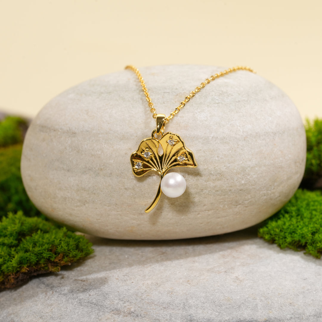 A product shot of a gold ginkgo leaf and pearl necklace elegantly draped over a smooth, light-colored stone. The stone is surrounded by clumps of bright green moss on a soft, neutral background.
