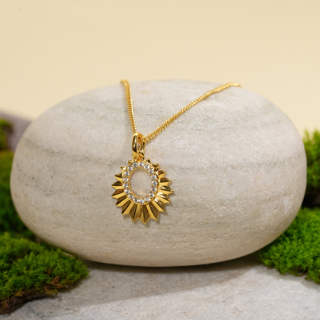 A beautiful gold sunburst necklace from Zewory Jewellery, displayed in a serene natural setting. The celestial pendant, with its faceted rays and crystal pavé center, is draped over a smooth, light-grey stone and surrounded by fresh green moss.