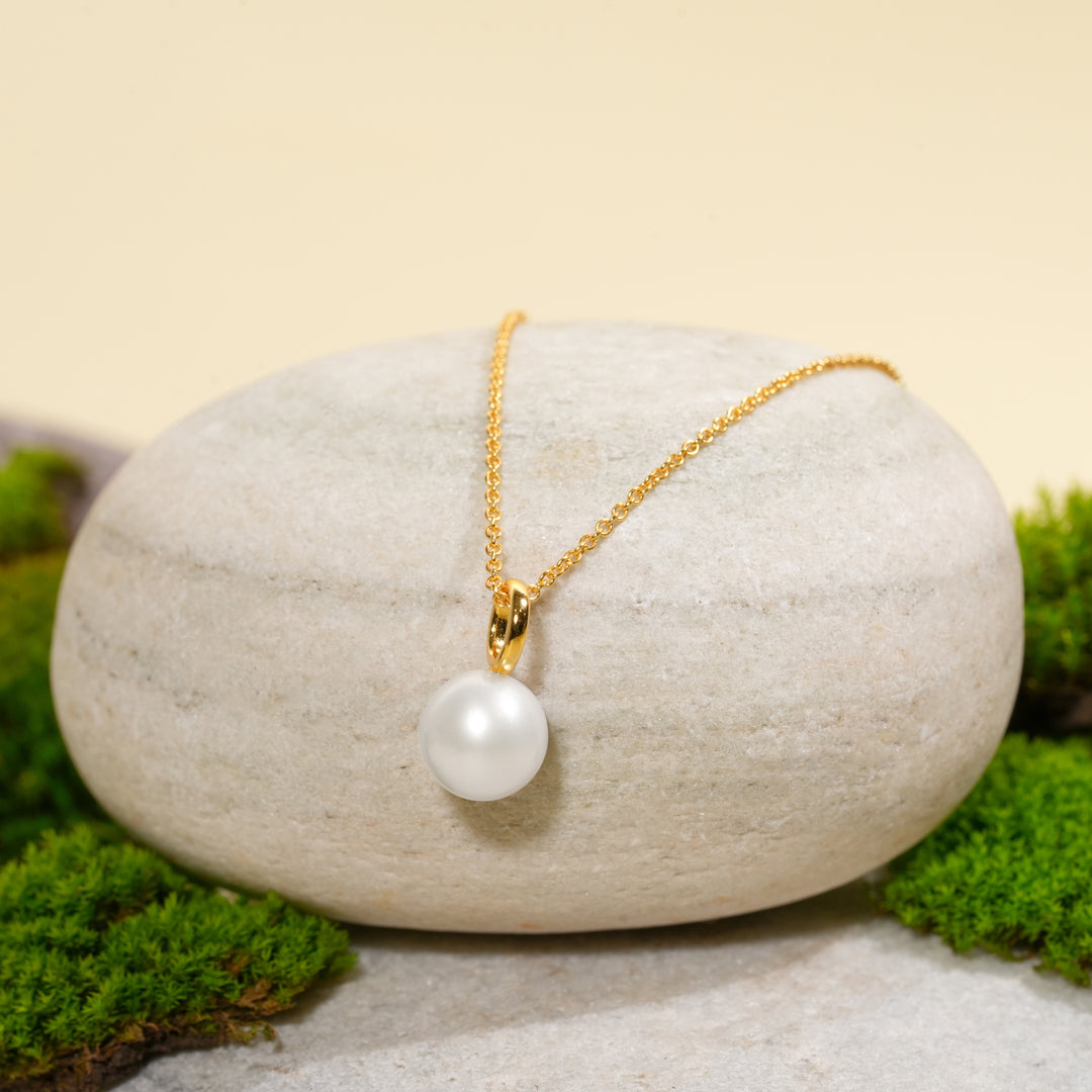 A gold necklace with a single, irregular pearl pendant is draped over a smooth, light-colored stone. The display is surrounded by vibrant green moss against a warm, neutral background.