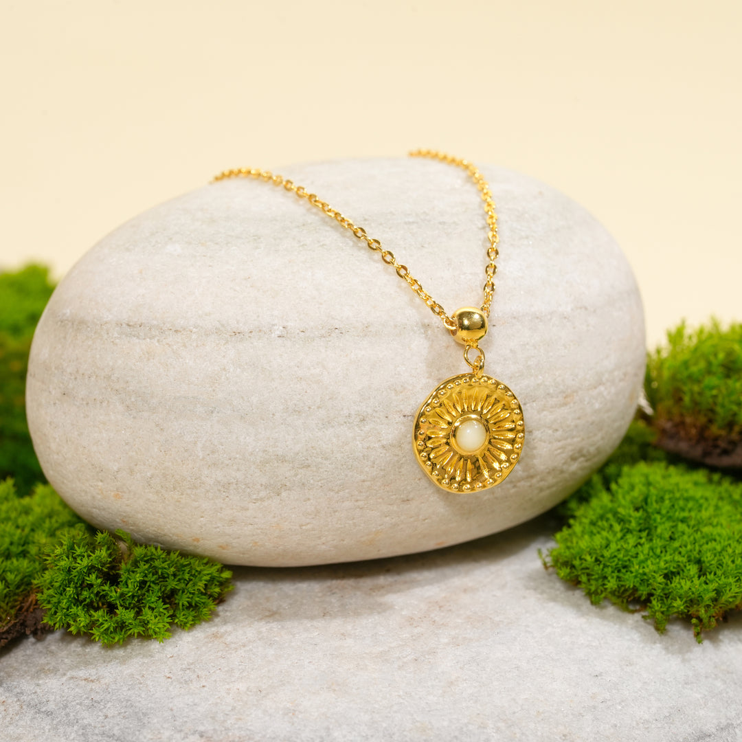 A beautiful gold sunburst necklace with a central opalescent stone, displayed in a serene natural setting. The celestial pendant from Zewory Jewellery is draped over a smooth, light-grey stone and surrounded by fresh green moss.