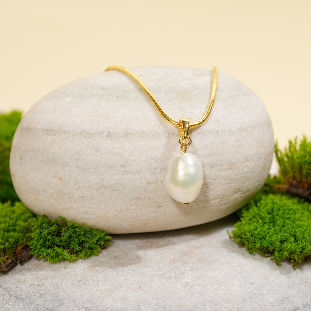 A gold necklace with a single, irregular pearl pendant is draped over a smooth, light-colored stone. The display is surrounded by vibrant green moss against a warm, neutral background.