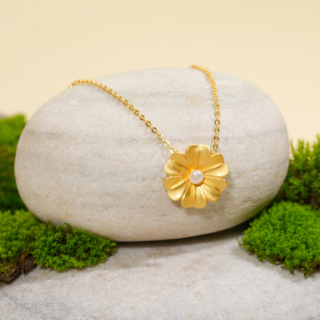 An elegant gold flower necklace with a pearl center, displayed in a serene, natural setting. The beautiful floral pendant from Zewory Jewellery is draped over a smooth, light-grey stone and surrounded by fresh green moss.