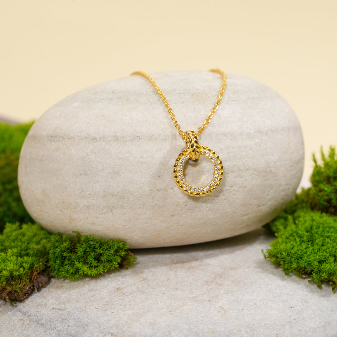 An elegant gold circle necklace from Zewory Jewellery, displayed in a serene natural setting. The sparkling pavé crystal and twisted rope-textured pendant is draped over a smooth, light-grey stone and surrounded by fresh green moss.