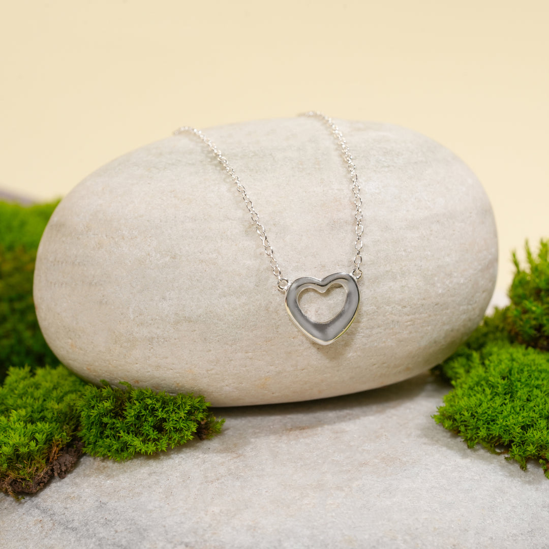 A delicate silver open heart necklace from Zewory Jewellery, displayed in a serene natural setting. The minimalist, polished heart pendant is draped over a smooth, light-grey stone and surrounded by fresh green moss.