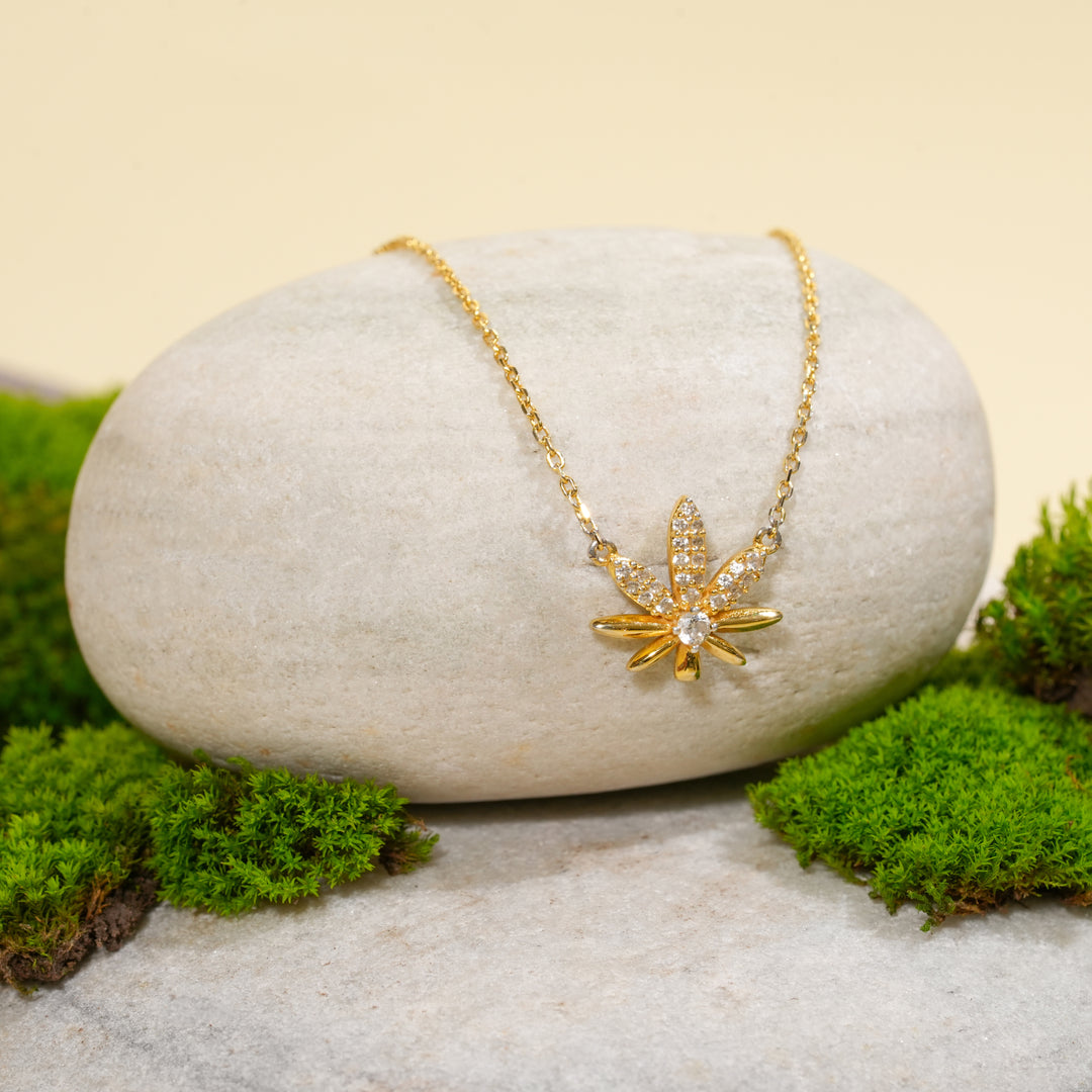An elegant gold cannabis leaf necklace from Zewory Jewellery, displayed in a serene natural setting. The sparkling, crystal-pavé pot leaf pendant is draped over a smooth, light-grey stone and surrounded by fresh green moss.