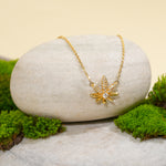 Load image into Gallery viewer, An elegant gold cannabis leaf necklace from Zewory Jewellery, displayed in a serene natural setting. The sparkling, crystal-pavé pot leaf pendant is draped over a smooth, light-grey stone and surrounded by fresh green moss.
