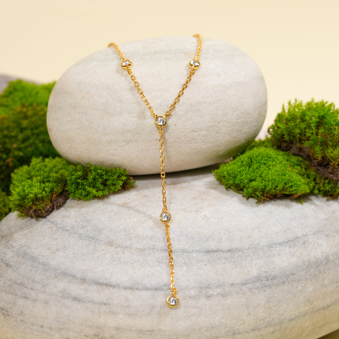 An elegant gold lariat necklace from Zewory Jewellery, displayed in a serene natural setting. The delicate Y-necklace, sparkling with bezel-set crystals, is draped over a smooth, light-grey stone and surrounded by fresh green moss.