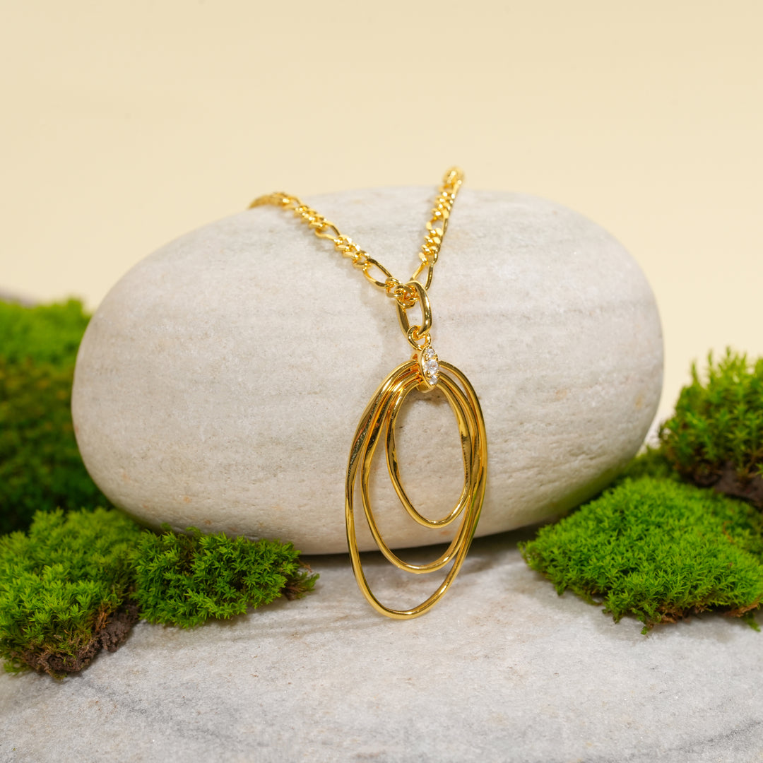 An elegant gold figaro chain necklace with an abstract, multi-loop oval pendant from Zewory Jewellery. The contemporary piece is displayed on a smooth, light-grey stone and surrounded by fresh green moss for a natural aesthetic.