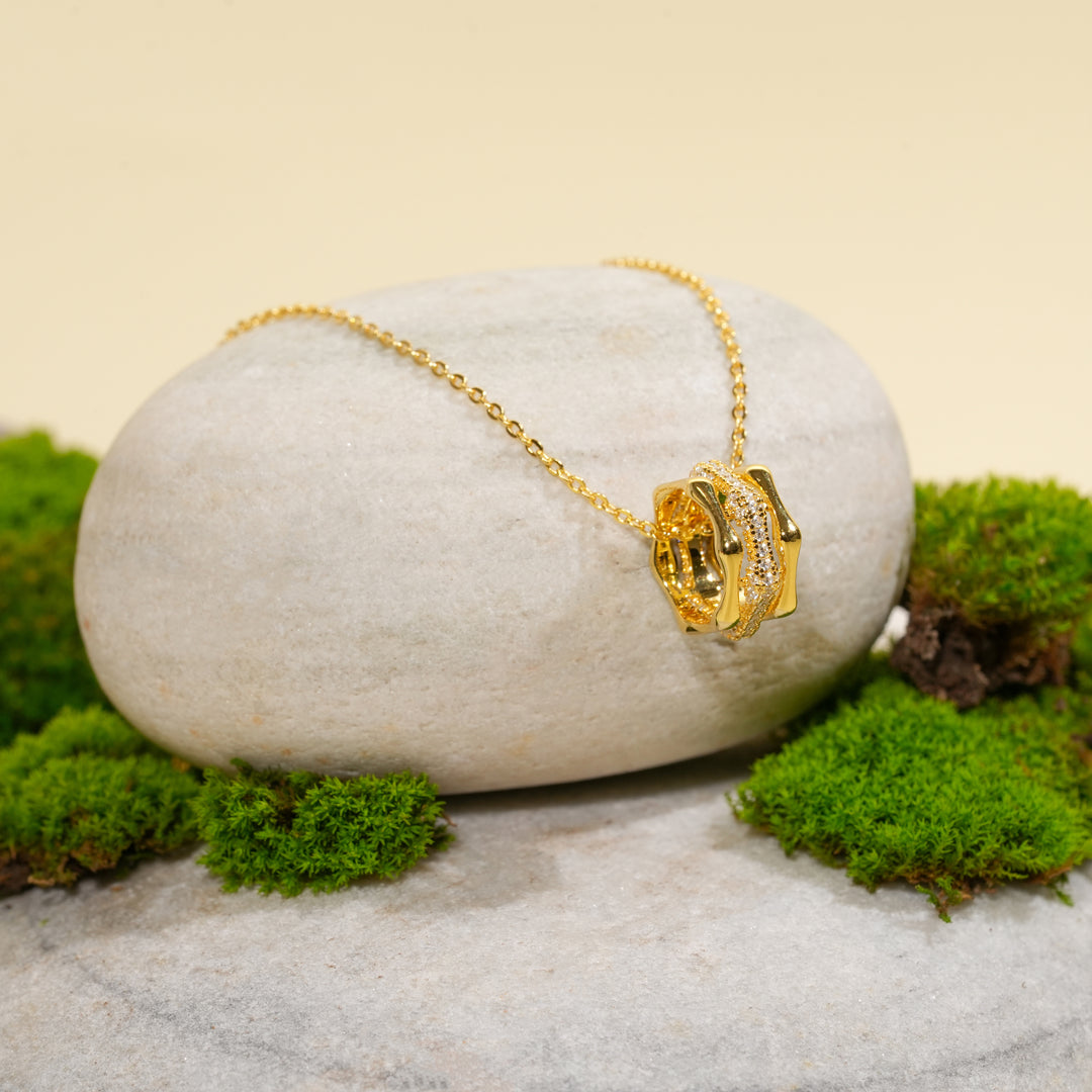 A contemporary gold necklace with a sparkling, crystal-accented barrel pendant from Zewory Jewellery. The elegant piece is displayed on a smooth, light-grey stone and surrounded by fresh green moss for a natural aesthetic.