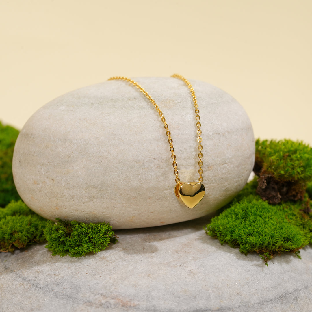 A delicate gold heart necklace from Zewory Jewellery, displayed in a serene natural setting. The minimalist, polished heart pendant is draped over a smooth, light-grey stone and surrounded by fresh green moss.