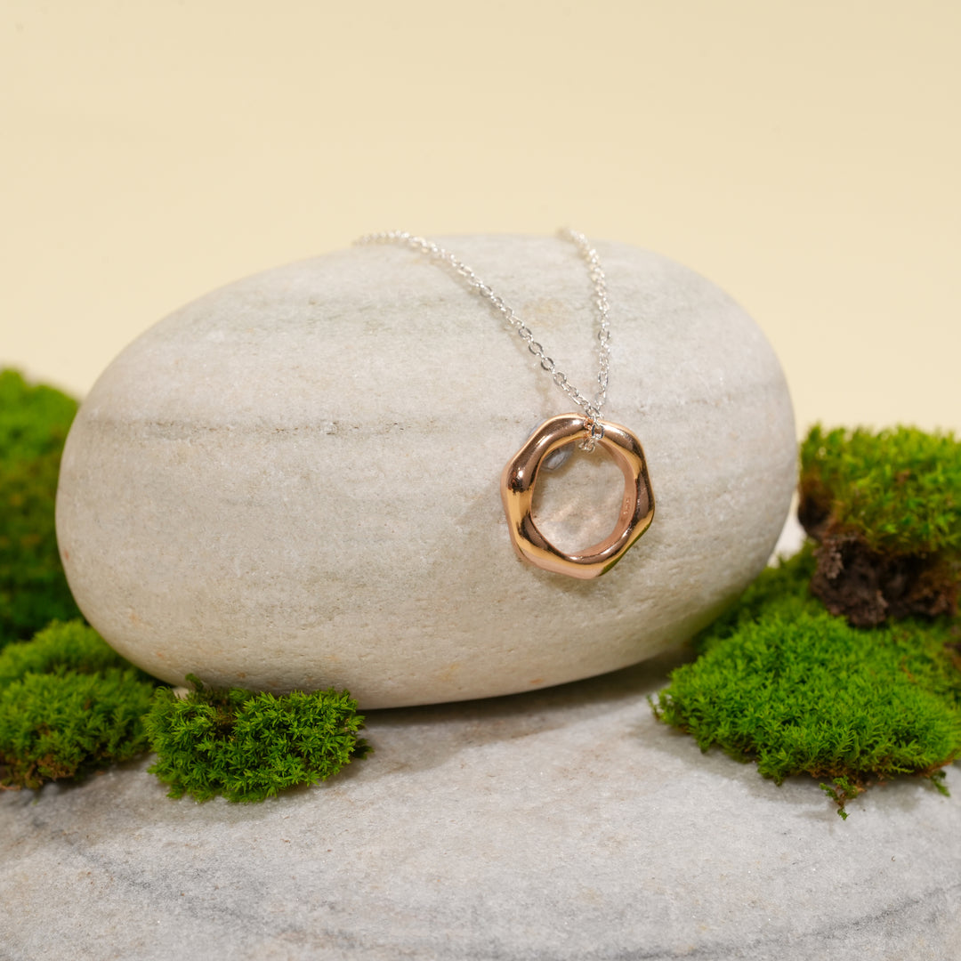 A contemporary two-tone necklace from Zewory Jewellery, displayed in a serene natural setting. The unique, molten-style rose gold circle pendant hangs from a silver chain, draped over a smooth, light-grey stone and surrounded by fresh green moss.